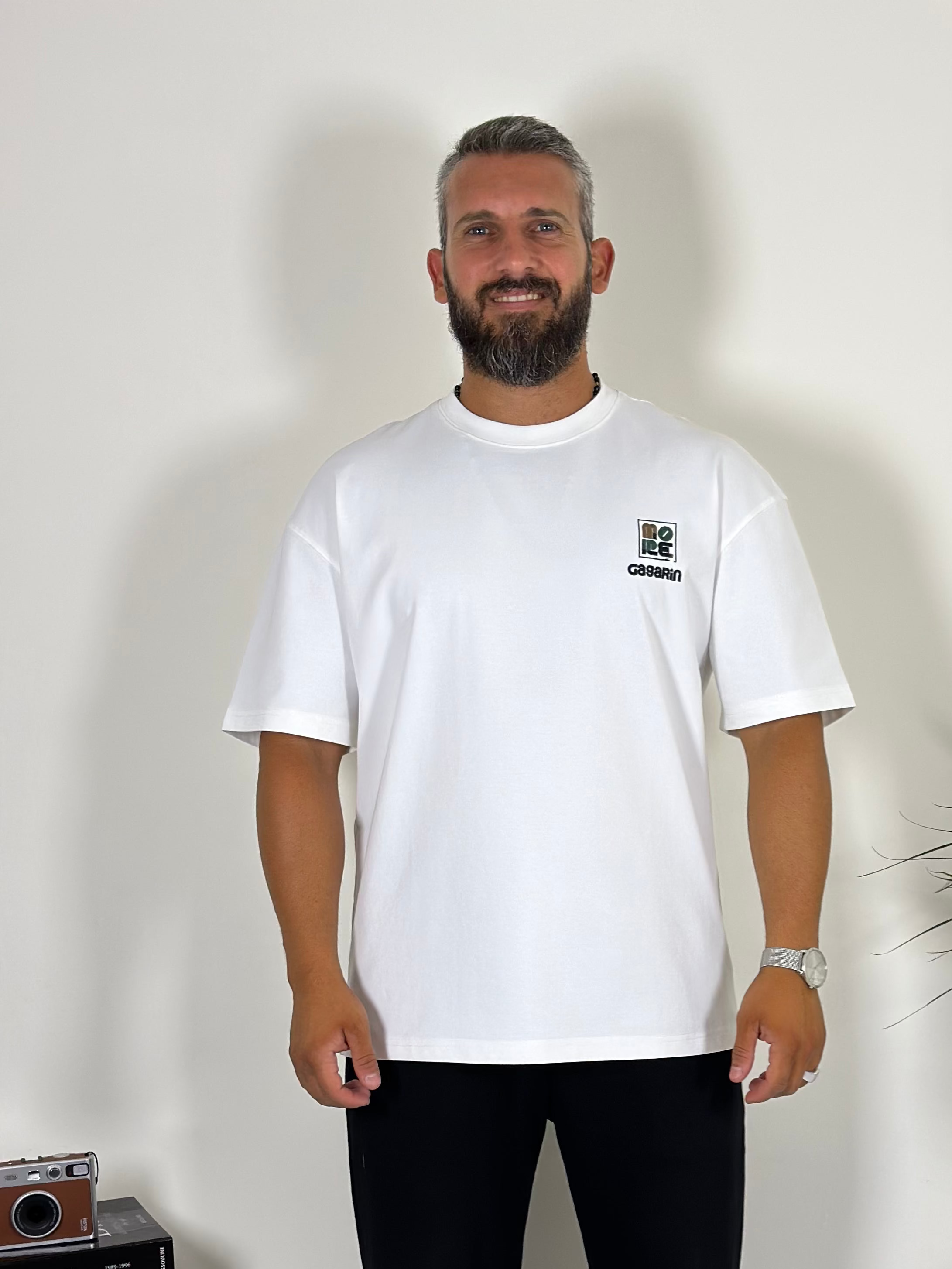 Man wearing a white t-shirt with a small logo, standing indoors next to a plant and a small table.
