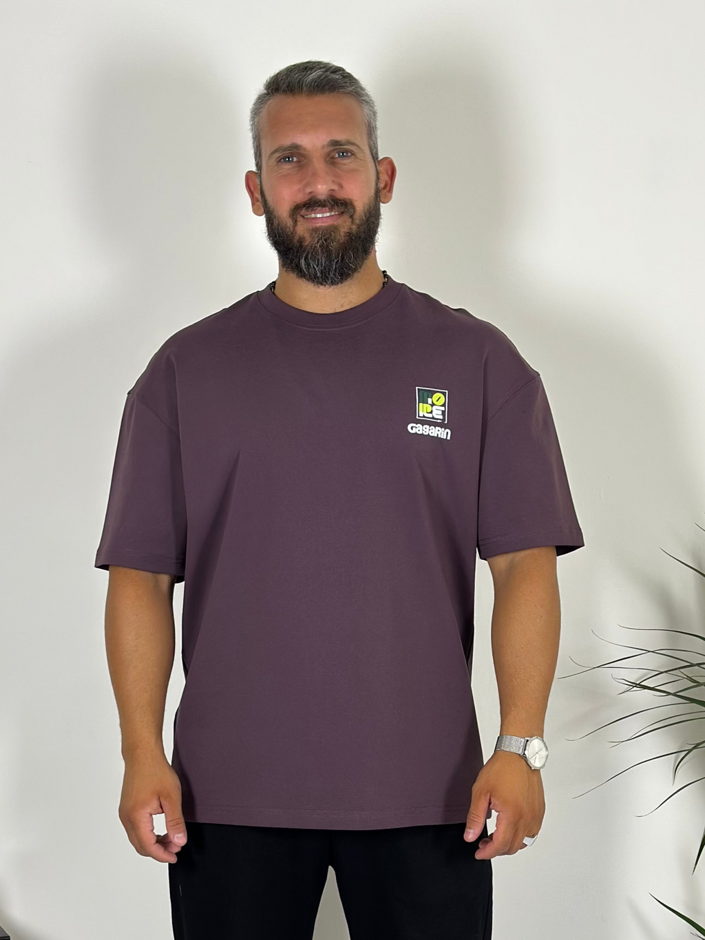 Man wearing a dark purple t-shirt with a logo, standing indoors next to a plant.