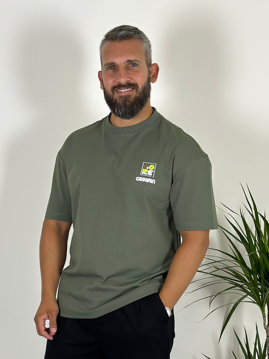 Man wearing a green t-shirt with a logo standing next to a plant indoors.