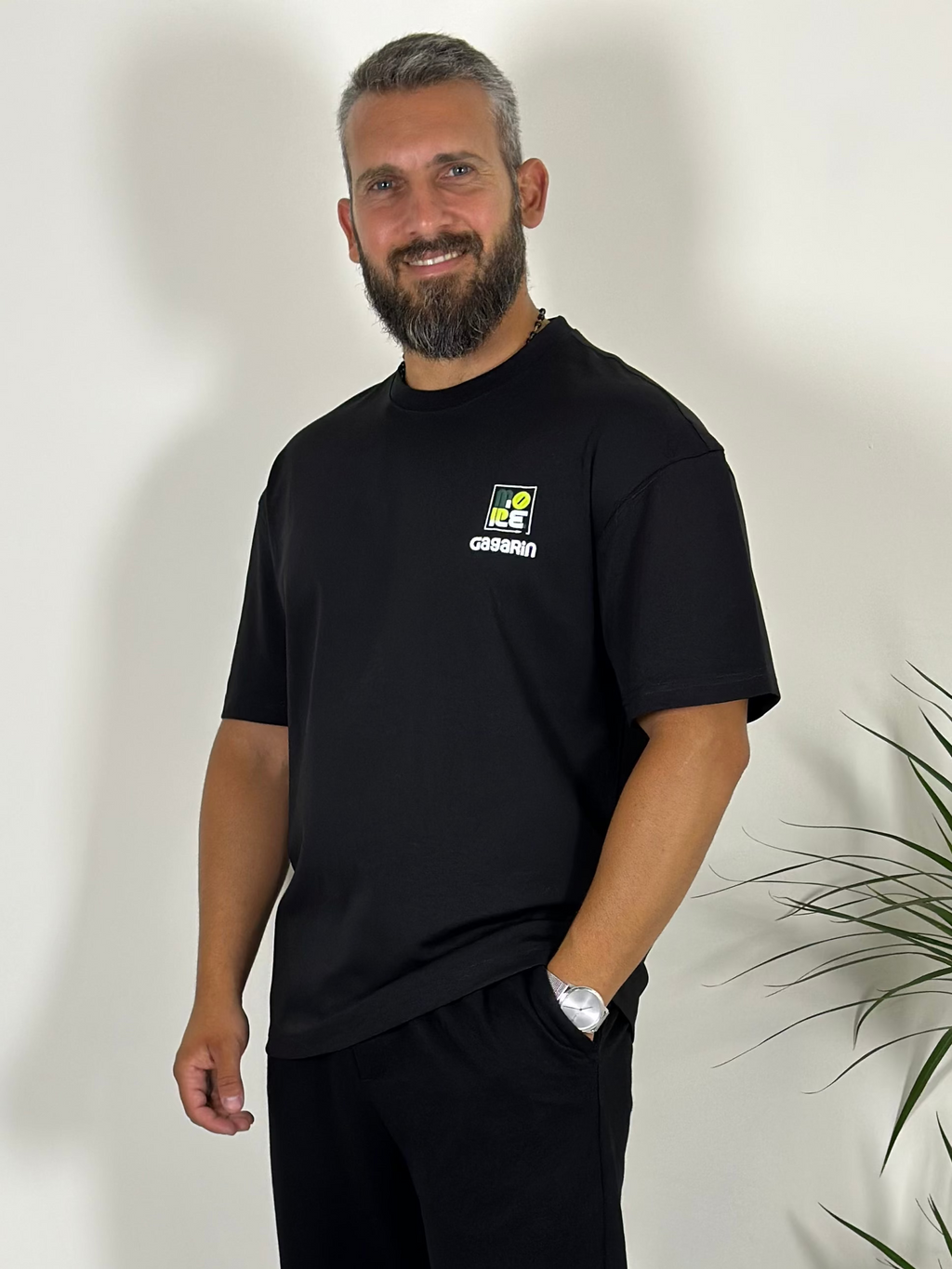 Man wearing a black t-shirt with a logo standing in a room with a plant and a shelf.