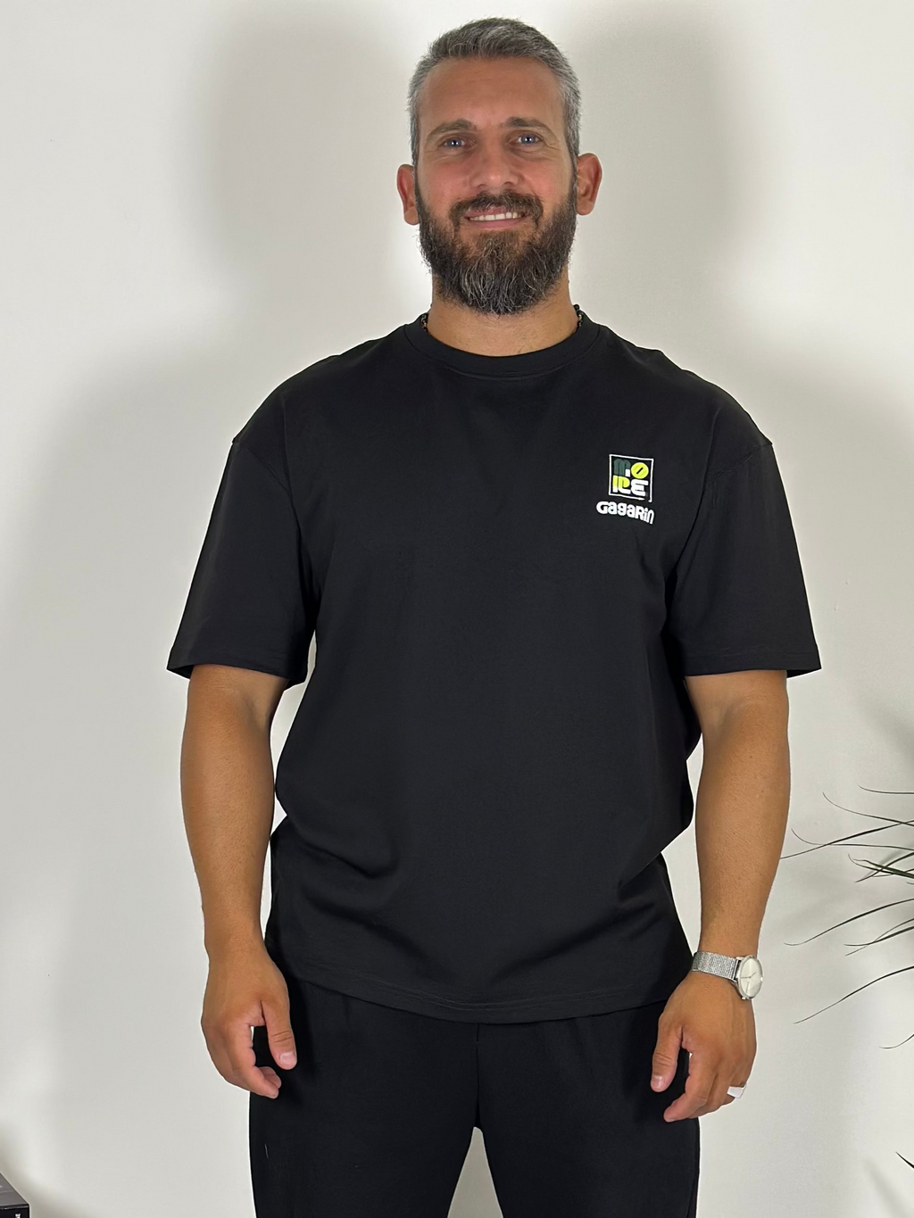 Man wearing a black t-shirt with a logo standing next to a plant indoors.