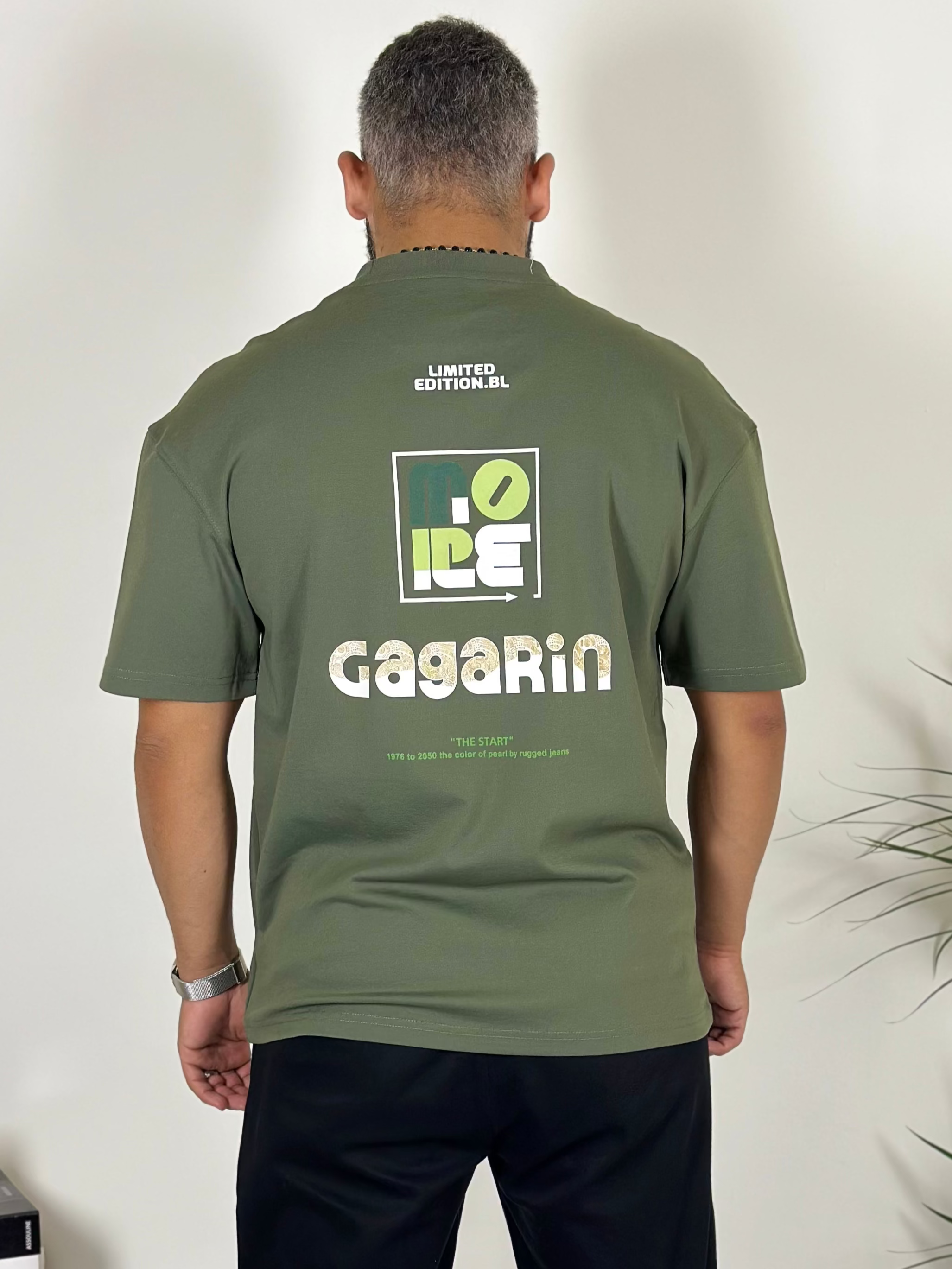 Person wearing a green t-shirt with branding, standing in a room with a plant.